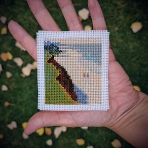 May include: A small, square cross-stitch landscape featuring a tree, a river, and a cloudy sky. The artwork is framed with a white border and held in a hand against a backdrop of green grass and fallen leaves. The color palette includes greens, blues, browns, and beige.