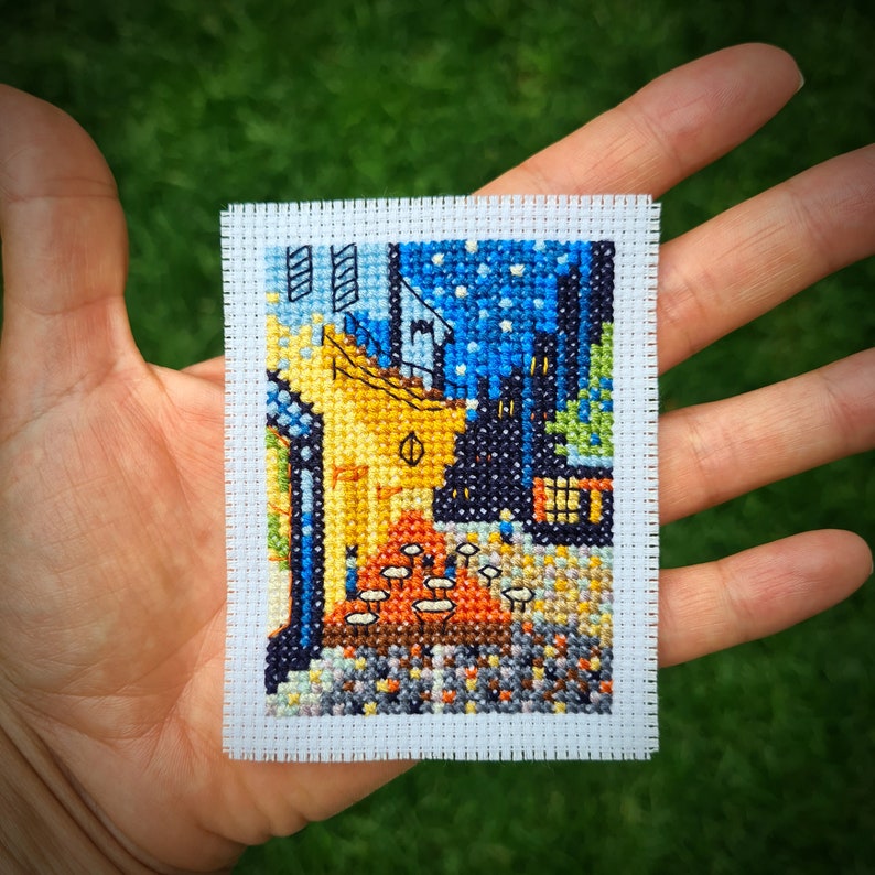 Puede incluir: Una bordado de punto de cruz de una pintura famosa que representa una escena nocturna con edificios y un cielo estrellado. El bordado est&aacute; enmarcado con un borde blanco y se sostiene en una mano.