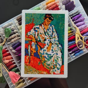 May include: A completed cross-stitch artwork depicting a figure in a colorful kimono, set against a green background. The piece is surrounded by a white border and rests on a tray of colorful embroidery floss. Gold scissors are also visible.