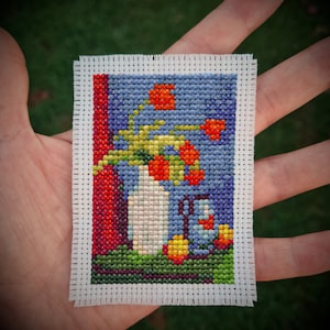 May include: A cross-stitch embroidery of a vase with a bouquet of red, orange, and yellow flowers. The vase is white and the background is blue. The embroidery is framed with a white border.