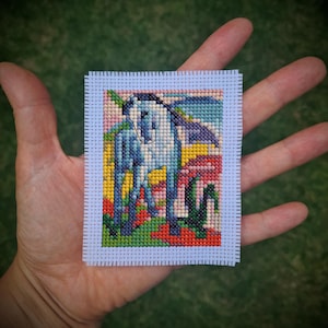 May include: A small, square cross-stitch artwork depicting a stylized horse in shades of blue, white, and black. The horse is set against a backdrop of colourful, abstract scenery with red, yellow, and green hues. The artwork has a light blue border.