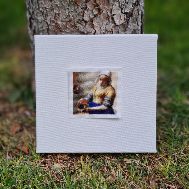 May include: A white canvas with a framed cross-stitch of a woman in a yellow dress and blue skirt holding a jug. The woman is standing in a room with a window and a lantern.