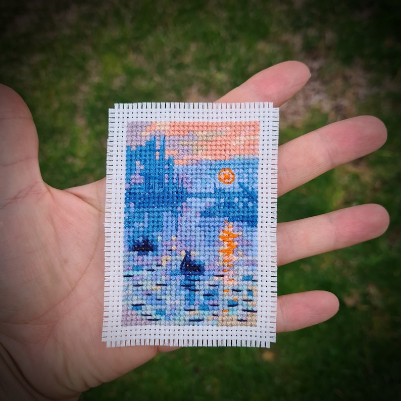 May include: A miniature cross-stitch landscape featuring a sunset over water. The scene includes a blue skyline, a bright orange sun, and reflections in the water. The artwork is framed with a white border and held in a hand.