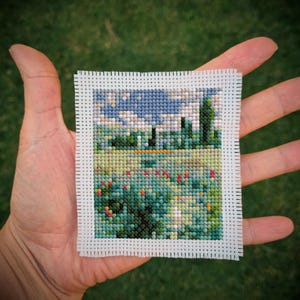 Può includere: Un piccolo paesaggio a punto croce con cielo blu, nuvole bianche, campi verdi e fiori colorati. L'opera è incorniciata con un bordo bianco ed è tenuta in una mano.