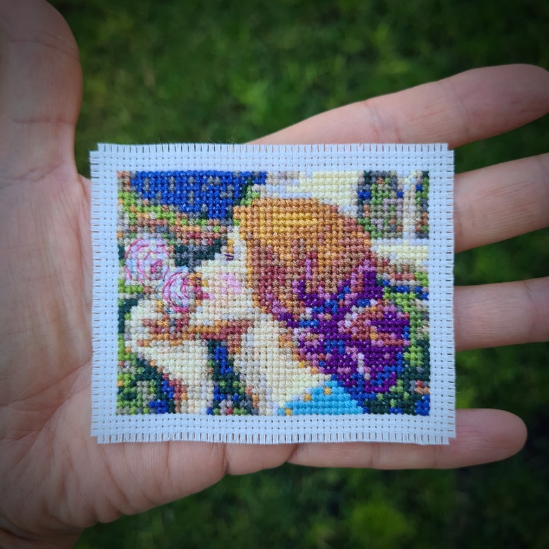 May include: A cross-stitch embroidery of two people with flowers and a blue background. The embroidery is framed with a white border.
