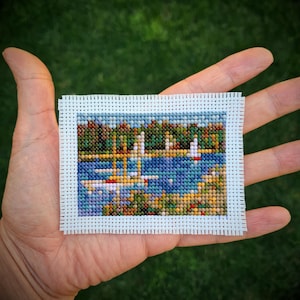 May include: A miniature cross-stitch landscape depicting a lake scene with sailboats and trees. The artwork features a blue lake, green and brown trees, and white sailboats. The cross-stitch is framed with a white border and held in a hand.