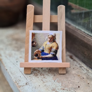 May include: A cross-stitch embroidery of a woman in a yellow dress and blue skirt, holding a jug of milk. The embroidery is framed and displayed on a wooden easel.