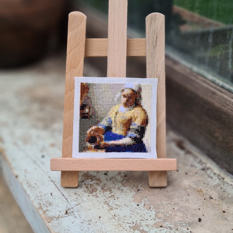 May include: A cross-stitch embroidery of a woman in a yellow dress and blue skirt, holding a jug of milk. The embroidery is framed and displayed on a wooden easel.