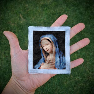Pode incluir: Uma pequena peça de ponto cruz quadrada que retrata uma figura religiosa com um véu azul e uma expressão serena. A obra de arte é emoldurada com uma borda branca e sobre um fundo preto. A peça é segurada em uma mão, mostrando os detalhes.