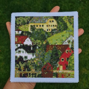 May include: A cross-stitch landscape scene featuring a village with yellow, red, and white buildings nestled among green trees. The artwork is framed with a light blue border and is held in a hand.