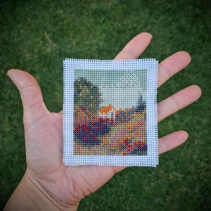 May include: A miniature cross-stitch picture showing a landscape with a house, trees, and flowers. The artwork is framed with white perforated fabric. The colour scheme includes greens, reds, oranges, and blues. The piece is held in a hand.