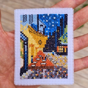 Puede incluir: Una bordado de punto de cruz que representa una pintura de un edificio amarillo con un techo azul, un cielo estrellado de noche y una escena de calle con peque&ntilde;as figuras. El bordado est&aacute; enmarcado con un borde blanco.