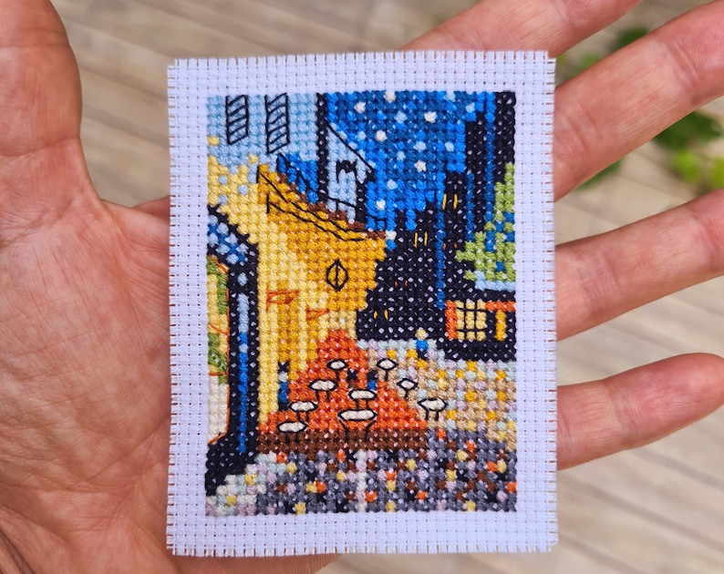 Puede incluir: Una bordado de punto de cruz que representa una pintura de un edificio amarillo con un techo azul, un cielo estrellado de noche y una escena de calle con peque&ntilde;as figuras. El bordado est&aacute; enmarcado con un borde blanco.