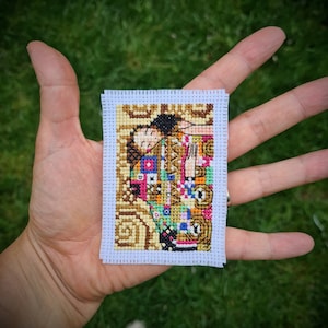 Può includere: Opera di ricamo a punto croce che rappresenta una coppia che si abbraccia, con uno sfondo a motivi geometrici colorati. L'opera è incorniciata da un bordo bianco con un passepartout bianco.