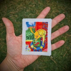 May include: A colorful cross-stitch pattern of a yellow cat sitting on a blue and green stool. The cat is surrounded by red, green, and blue geometric shapes.