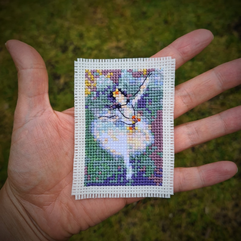 May include: A cross-stitch embroidery of a ballerina in a white dress with a colorful background.