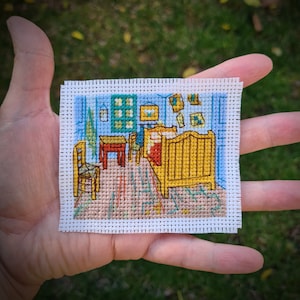 Puede incluir: Un pequeño punto de cruz que representa "La habitación en Arlés" de Van Gogh. La obra presenta una cama amarilla, una mesa roja y paredes azules. El punto de cruz está sobre tela blanca.