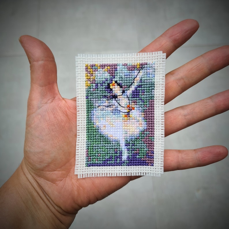 May include: A cross-stitch embroidery of a ballerina in a white tutu, with a colorful floral background. The embroidery is framed with a white border.