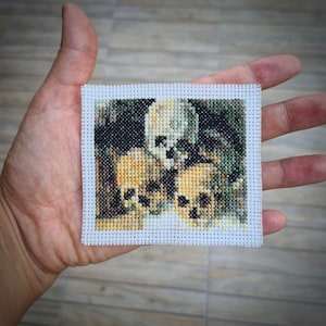 May include: A white cross-stitch embroidery with a design of three skulls with a green and brown background.