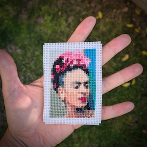 Puede incluir: Un bordado de punto de cruz de Frida Kahlo, una famosa pintora mexicana, con una corona de flores de colores y un fondo azul.