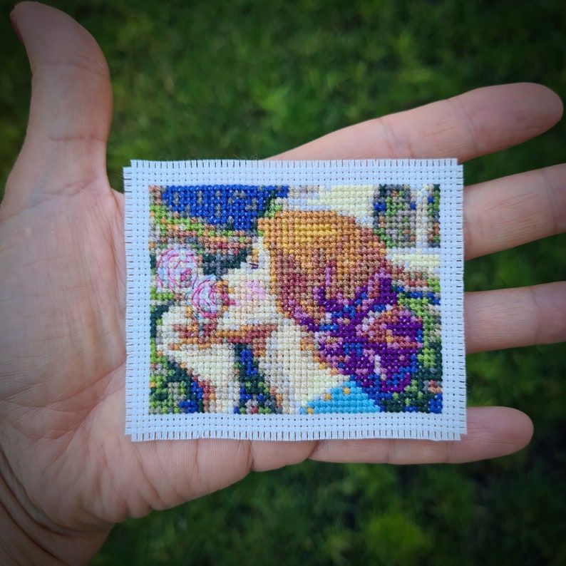 May include: A cross-stitch embroidery of a woman with blonde hair, wearing a blue dress, surrounded by pink and purple flowers.