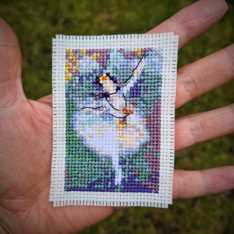 May include: A cross-stitch embroidery of a ballerina in a white tutu, with a green, purple, and yellow background.