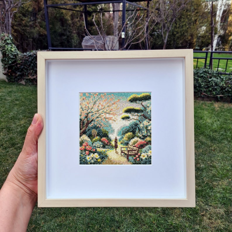 May include: A framed cross-stitch artwork depicting a serene garden scene with a path, trees, and flowers. The artwork is in a light wood frame with a white mat. The scene features a variety of colors, including greens, blues, and pinks.