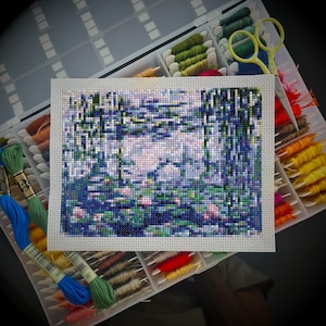May include: A completed cross-stitch project depicting a water garden scene with blue, green, and pink hues. The needlework is displayed in a white frame, surrounded by spools of colorful embroidery floss and a pair of yellow scissors in a clear organizer.