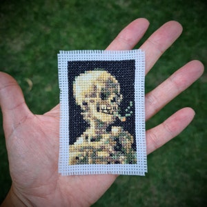 May include: A cross-stitch embroidery of a skeleton with a cigarette in its mouth. The skeleton is wearing a green and brown patterned shirt. The embroidery is framed with a white border.