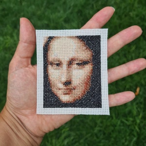 May include: A cross-stitch embroidery of a person's face. The face is rendered in shades of brown and white, with a black background.