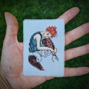 May include: A small, hand-stitched cross-stitch art piece featuring a stylized figure with orange hair and a blue vest, embracing a cluster of flowers. The artwork is on white Aida cloth, showcasing detailed needlework.