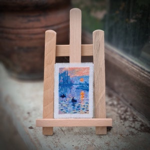 May include: A miniature cross-stitch landscape art piece depicting a sunset over water, displayed on a small wooden easel. The artwork features blue, orange, and purple hues. The easel is made of light-colored wood.