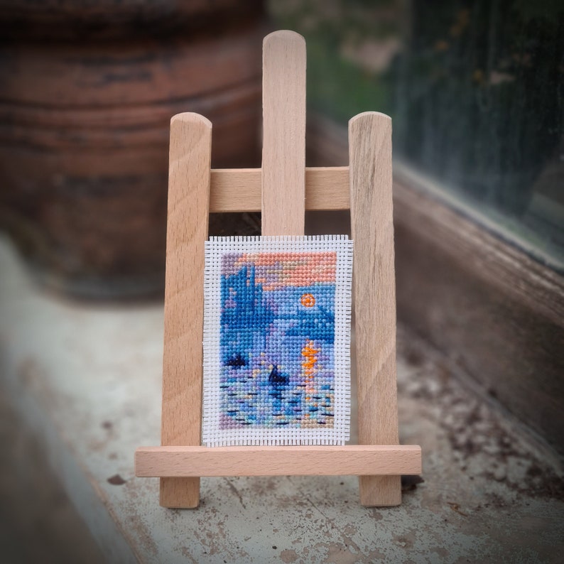 May include: A miniature cross-stitch landscape art piece depicting a sunset over water, displayed on a small wooden easel. The artwork features blue, orange, and purple hues. The easel is made of light-colored wood.