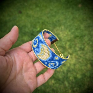 May include: A woven cuff bracelet featuring a blue and gold celestial pattern with a crescent moon and swirling star motifs. The bracelet is finished with a gold-toned metal chain and clasp mechanism, showcasing intricate beadwork and detailed stitching.