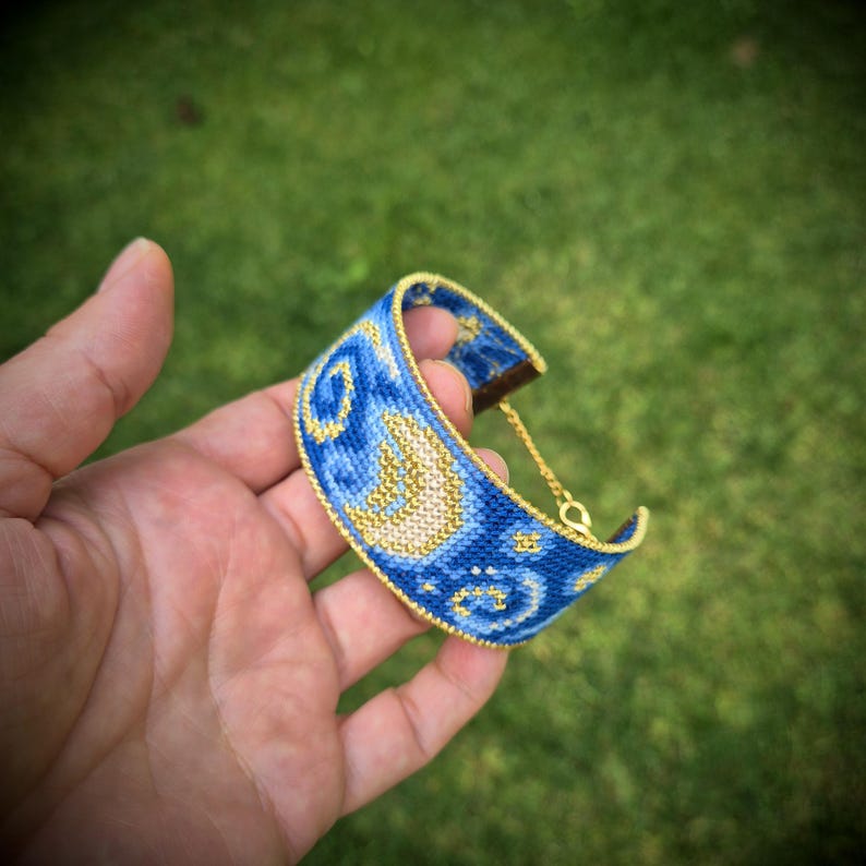 May include: A woven cuff bracelet featuring a blue and gold celestial pattern with a crescent moon and swirling star motifs. The bracelet is finished with a gold-toned metal chain and clasp mechanism, showcasing intricate beadwork and detailed stitching.