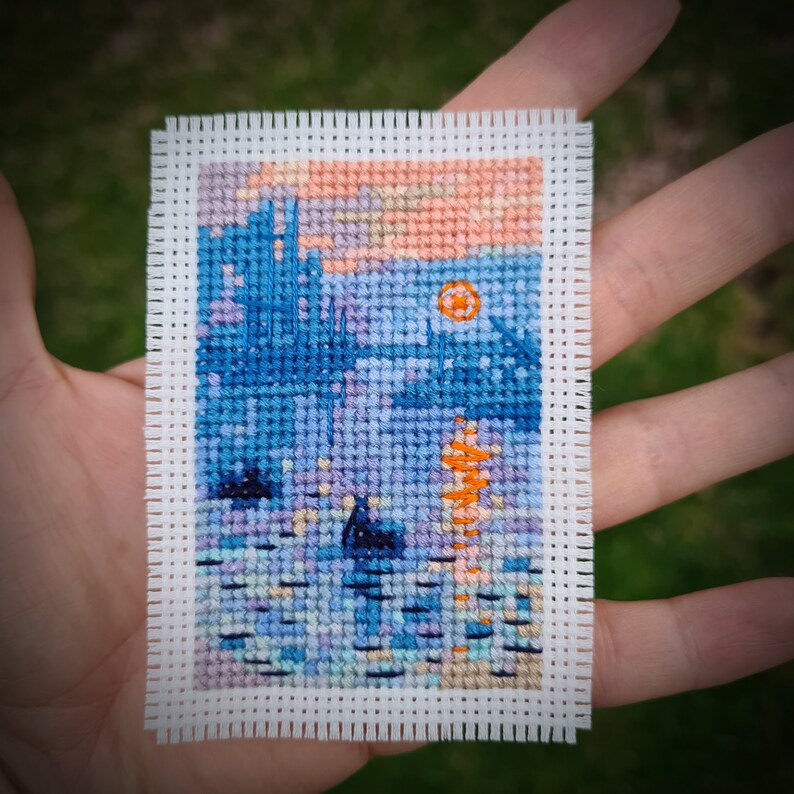 May include: A miniature cross-stitch landscape featuring a sunset over water. The scene includes a blue silhouette of a building, water reflections, and an orange sun. The artwork is on white fabric.