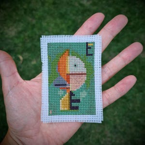 Op de afbeelding: Een klein, rechthoekig kruissteekstuk met een abstract ontwerp en de letter "E". Het ontwerp gebruikt oranje, geel en grijs tegen een groene achtergrond. Het heeft een witte rand.