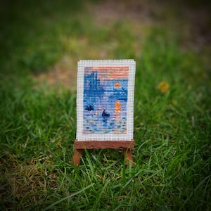 May include: A miniature cross-stitch artwork depicting a waterscape scene with a sunset, displayed on a small wooden easel. The artwork features shades of blue, orange, and yellow, with a detailed, textured appearance.