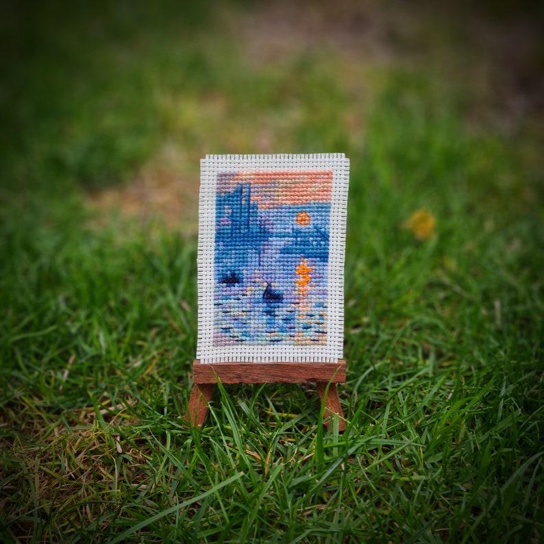 May include: A miniature cross-stitch artwork depicting a waterscape scene with a sunset, displayed on a small wooden easel. The artwork features shades of blue, orange, and yellow, with a detailed, textured appearance.
