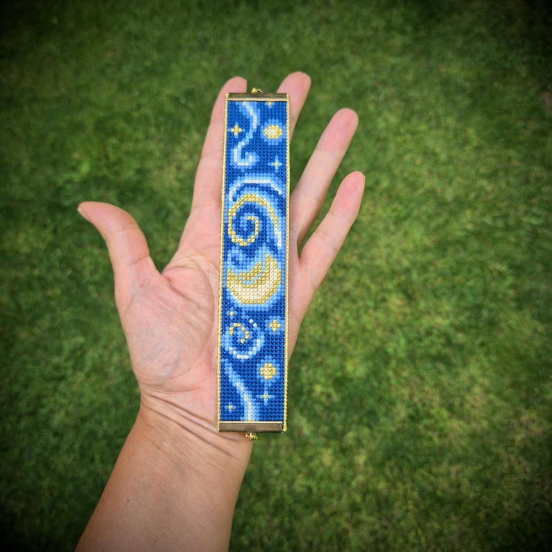 May include: A rectangular beaded bracelet featuring a blue background with a yellow crescent moon and star pattern. The design is composed of small, uniform beads arranged in a grid, framed by thin gold-toned metal edges at the top and bottom.