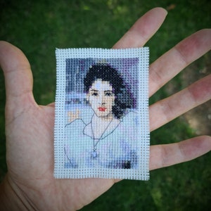 May include: A cross-stitch portrait of a woman with dark hair and a white shirt. The woman is wearing a silver necklace with a pendant. The portrait is framed in a white fabric.