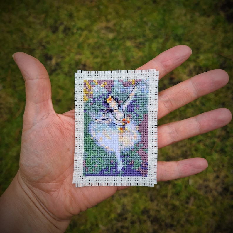 May include: A cross-stitch embroidery of a ballerina in a white tutu, with a green and purple background.