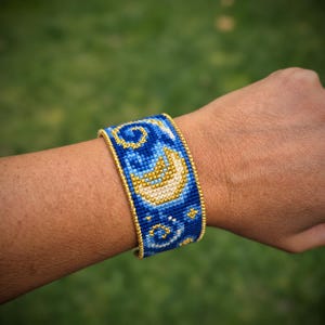 May include: A wide woven bracelet featuring a blue background with a repeating pattern of yellow crescent moons and swirling star motifs. The edges of the bracelet are finished with a gold-colored beaded trim that runs along the top and bottom.