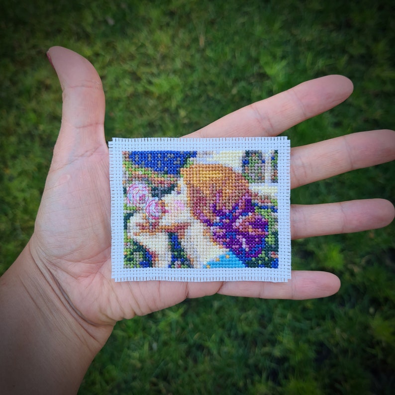 May include: A cross-stitch embroidery of a woman with brown hair smelling a purple flower. The background is a green and blue landscape.