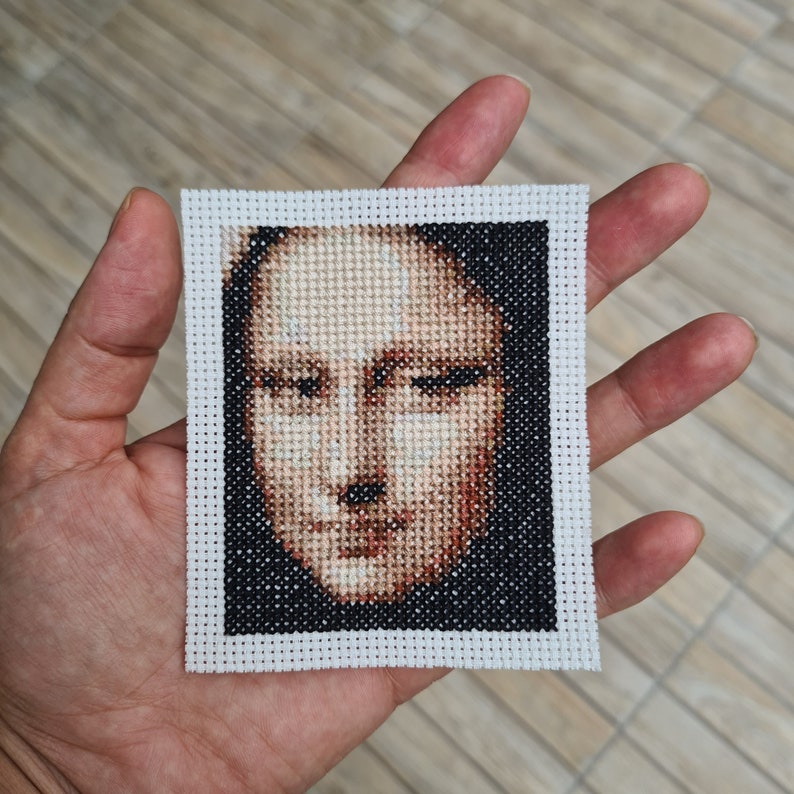 May include: A close-up of a cross-stitch embroidery of a person's face. The face is rendered in shades of brown, white, and black. The embroidery is framed with a white border.