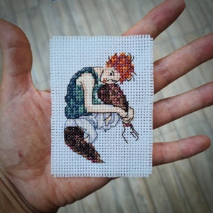 May include: A small, square cross-stitch depicting a figure with orange hair, a green vest, and white pants. The figure is hunched over, hugging a brown object. The cross-stitch is on a white fabric background.