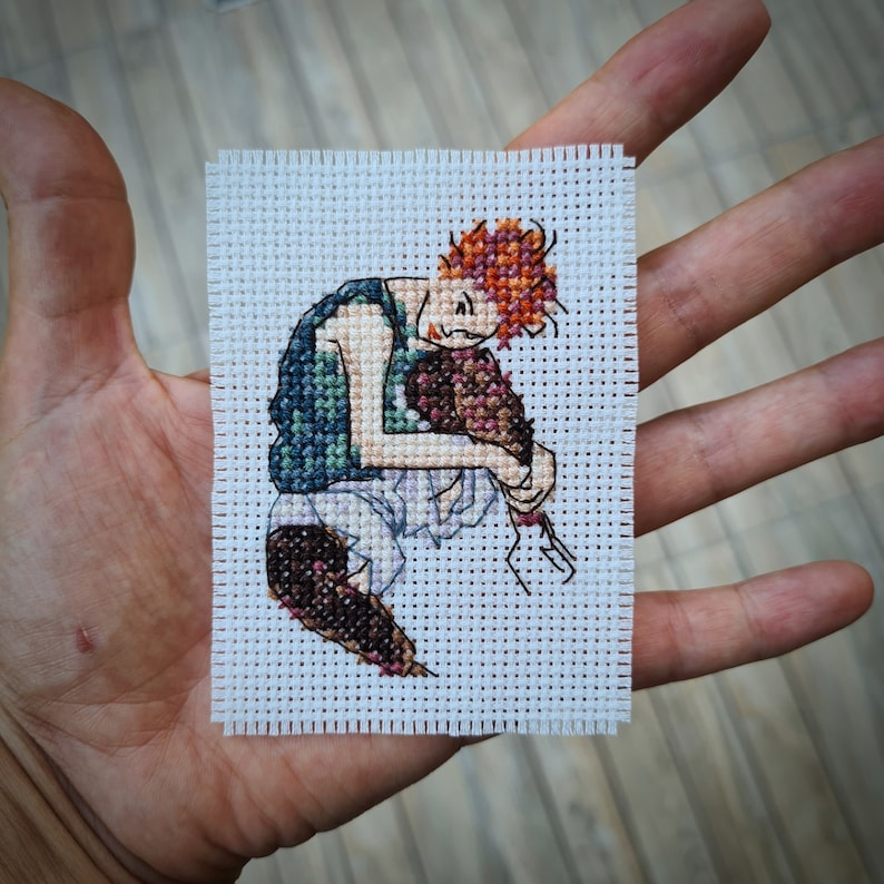 May include: A small, square cross-stitch depicting a figure with orange hair, a green vest, and white pants. The figure is hunched over, hugging a brown object. The cross-stitch is on a white fabric background.