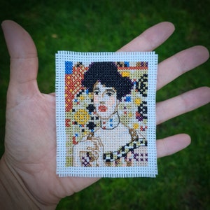 Può includere: Ricamo a punto croce di una donna con capelli scuri su uno sfondo colorato. Indossa un vestito bianco con un motivo bianco e nero.