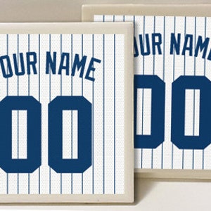 May include: Two white ceramic coasters with a blue and white baseball jersey design. The coasters feature a white background with blue vertical stripes and a blue number "00" with the text "YOUR NAME" above the number.