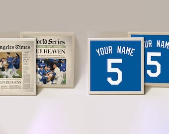 May include: Two blue and white ceramic coasters with the text "YOUR NAME" and the number "5" in white. The coasters are next to two newspaper clippings, one from the Los Angeles Times with the headline "THE REIGN RETURNS" and the other from the World Series with the headline "BLUE HEAVEN".